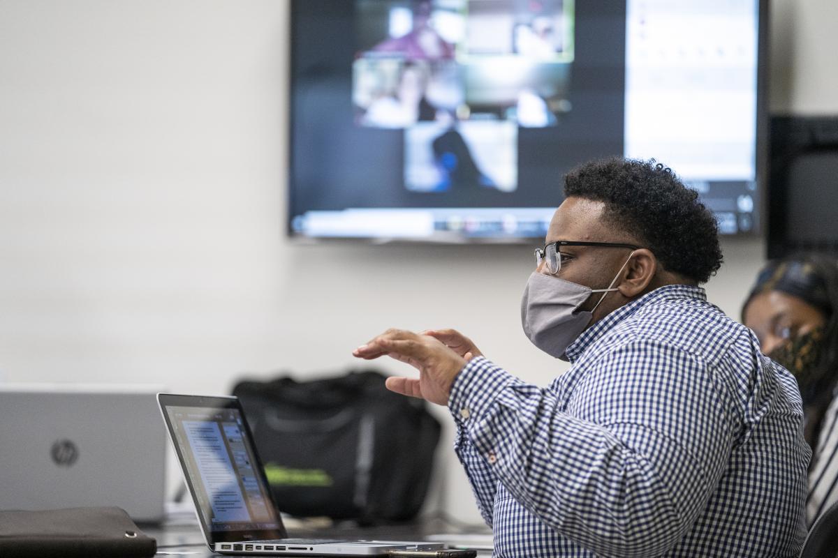 Man with a face mask talking in front of a screen